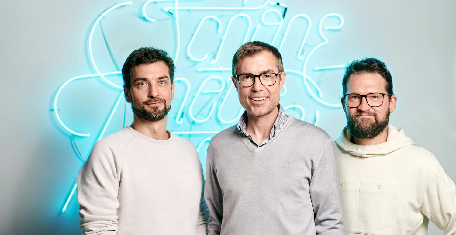 A picture of three men standing next to each other in front of an SNK neon sign in the SNK office